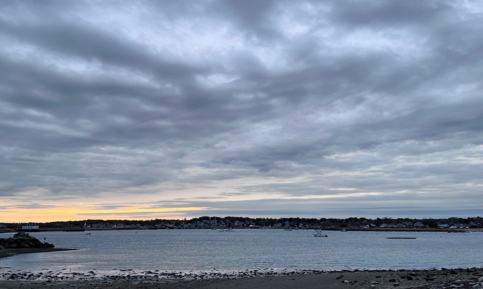 Grey clouds hang over harbor, the water is calm, it is winter, only a few boats remain moored in the harbor. The sky is clear on distant horizon, it turns yellow and organs with the setting sun.