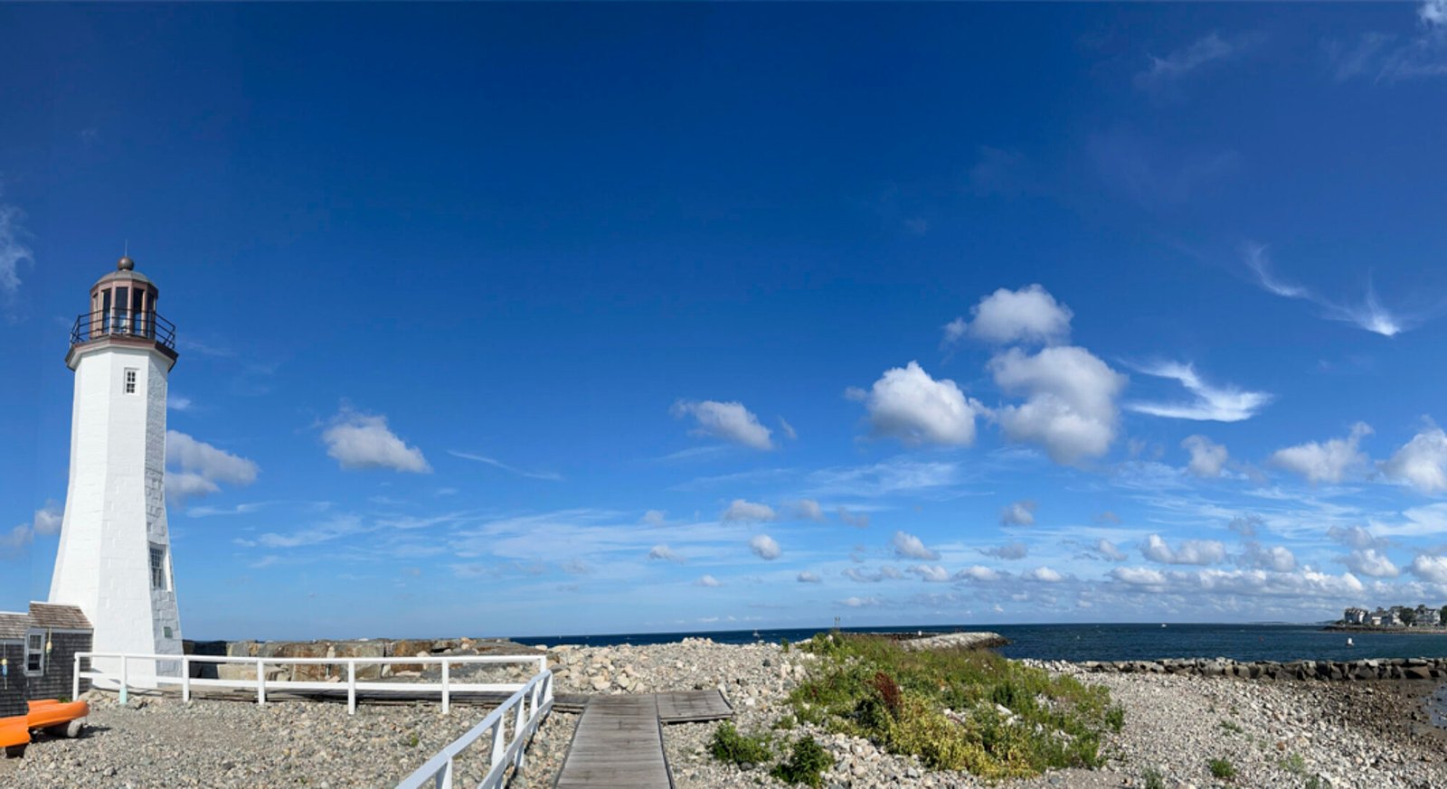 A blue sky with puffy white clouds hangs over a rocky shore. A bright white lighthouse stands in the left of the image, a wooden walkway leads up to its base, and seawall made of huge granite blocks stretches into the mouth of the harbor. The water is blue-green, and the sun is shining brightly in the west, over the harbor and town of Scituate, Massachusetts.